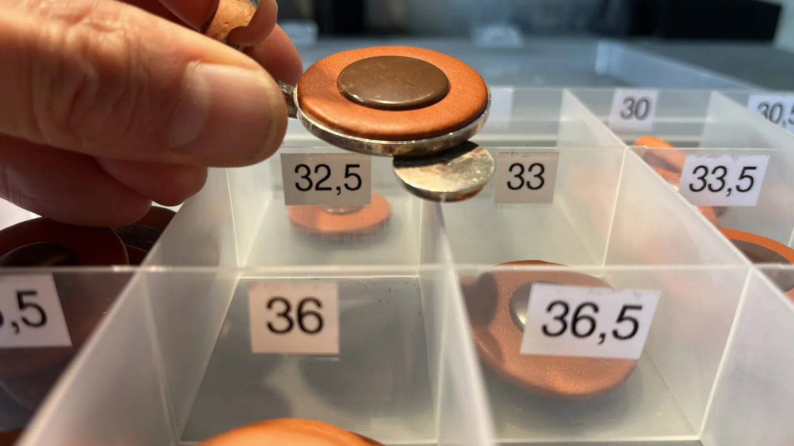 A saxophone key with an orange pad held up over assortment boxes with pads sorted by diameter, labelled 32.5, 33, 33.5, 36 and 36.5